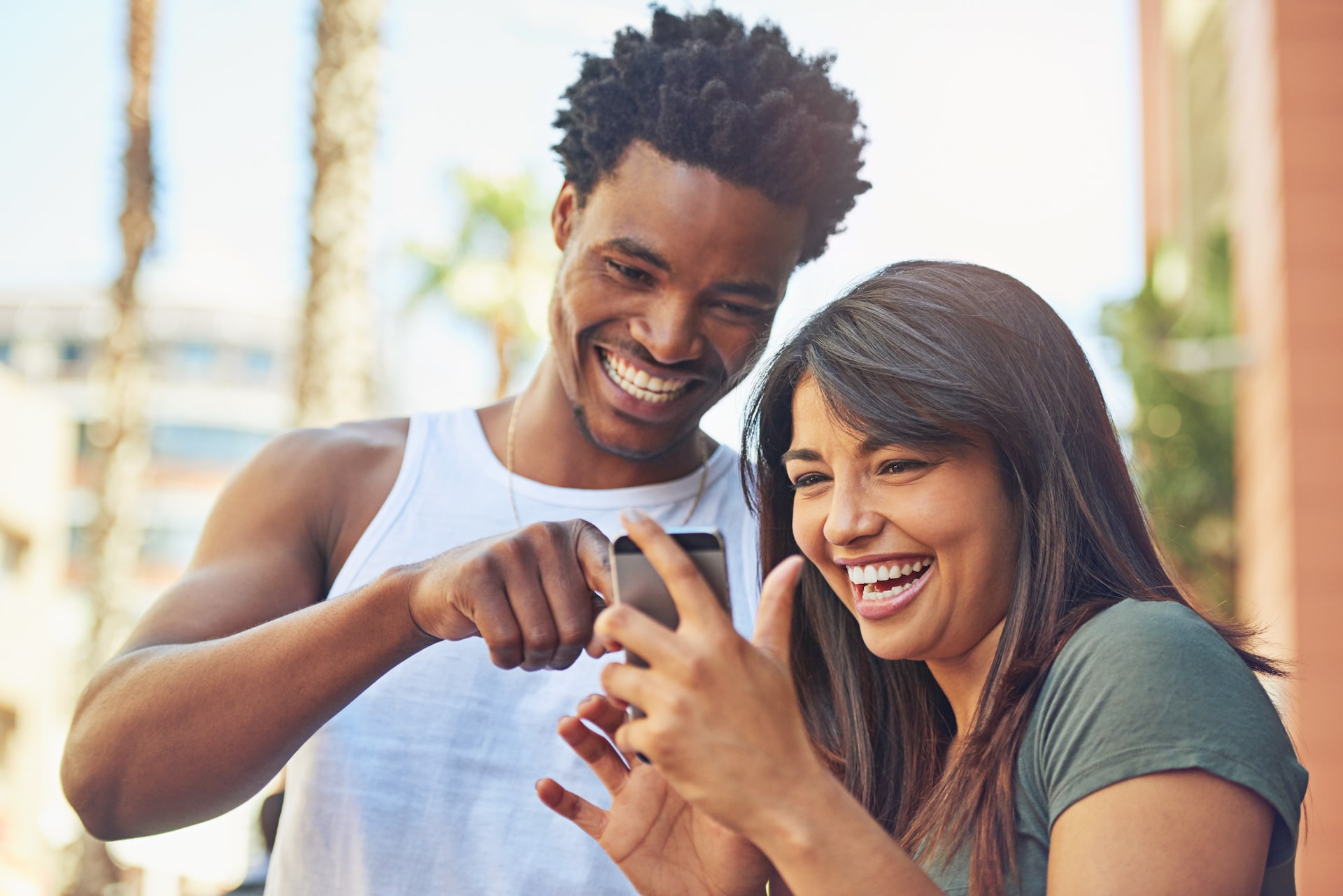 Two people, a man and women, looking at a phone and laughing. They are outside.
