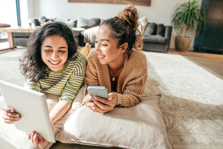 Two students lying on pillows on the floor with digital tablet and cellphone.